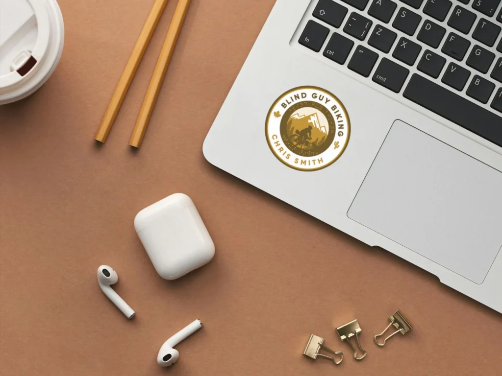 Looking down on a brown table, a Blind Guy Biking sticker is shown on the corner of a laptop keyboard. Also shown are Airpods, binder clips, and two unsharpened pencils.